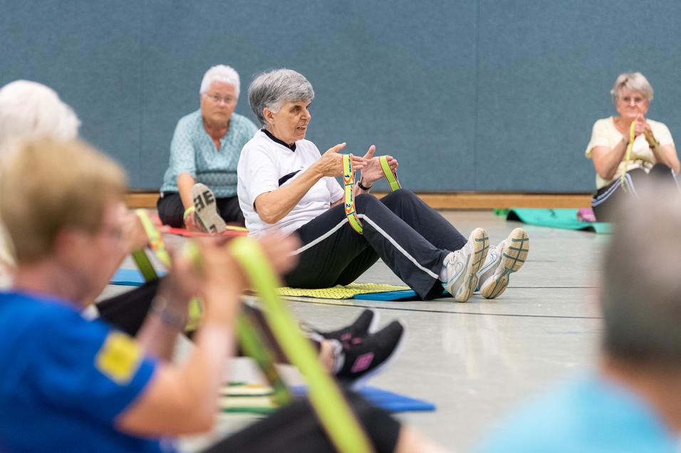 Senioren nehmen an einer Fitnessübung mit Fitnessbändern in einer Sporthalle teil, sitzend auf Fitnessmatten.