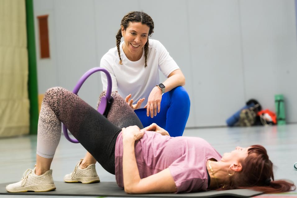 Zwei Frauen in Sportkleidung trainieren auf einer Matte, eine unterstützt die andere mit einem Fitnessgerät.