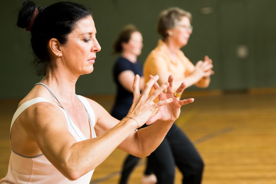Drei Frauen führen eine Fitnessübung in einem Saal mit Holzfußboden und einer Sporttorwand im Hintergrund aus.