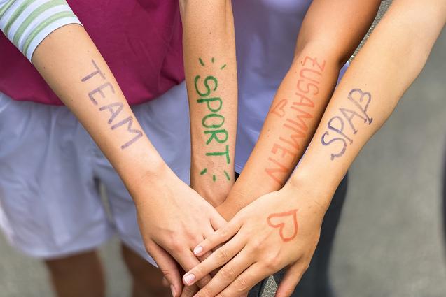 Vier Arme liegen übereinander, mit farbiger Schrift: „TEAM“, „SPORT“, „ZUSAMMEN“, „SPASS“ auf der Haut.