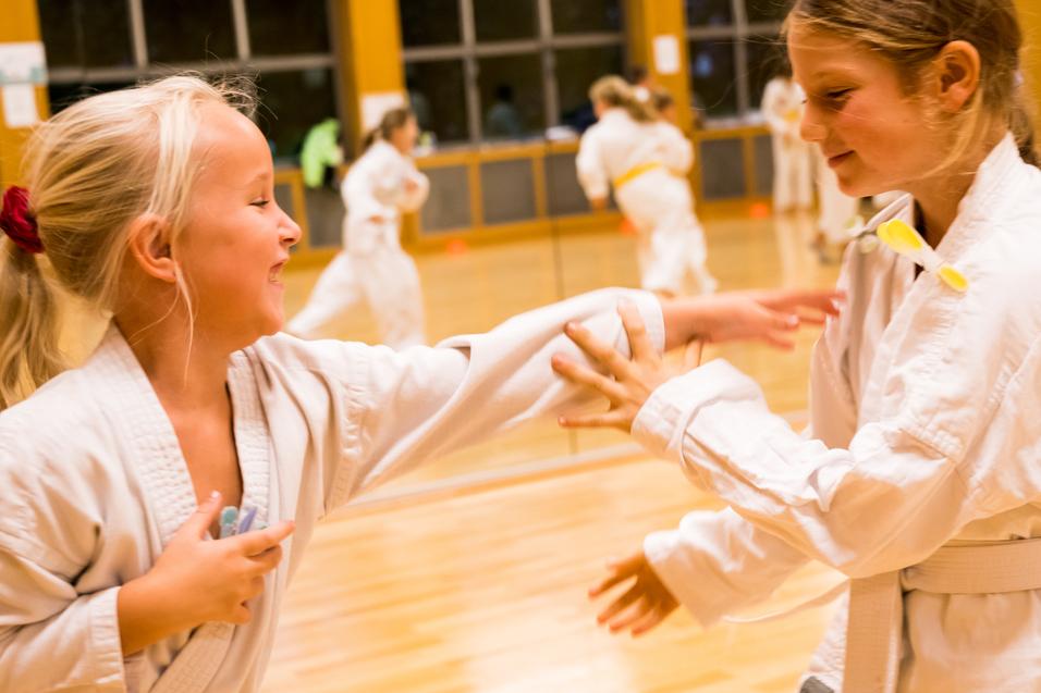 Zwei Mädchen im Karateanzug üben spielerisch im Trainingsraum miteinander, umgeben von weiteren Trainierenden.