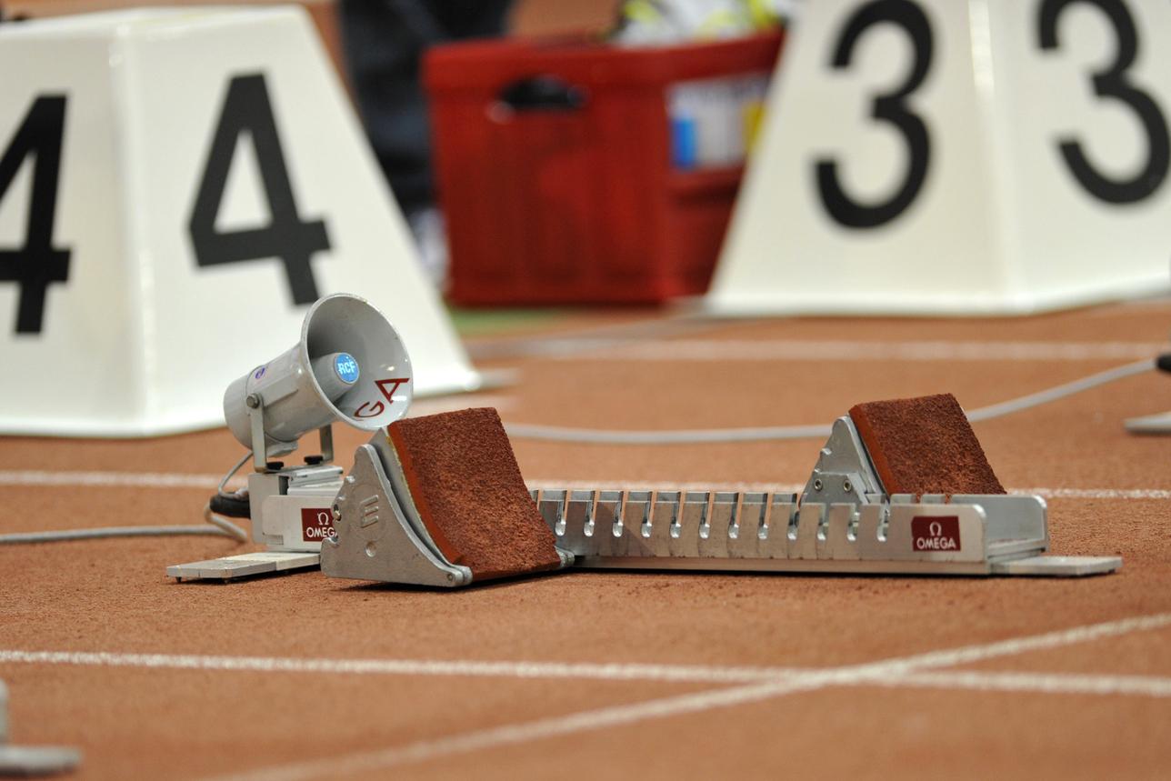 Startblöcke und Zeitmessgeräte auf einer Leichtathletikbahn mit brauner Oberfläche und großen Wettkampfnummern im Hintergrund.