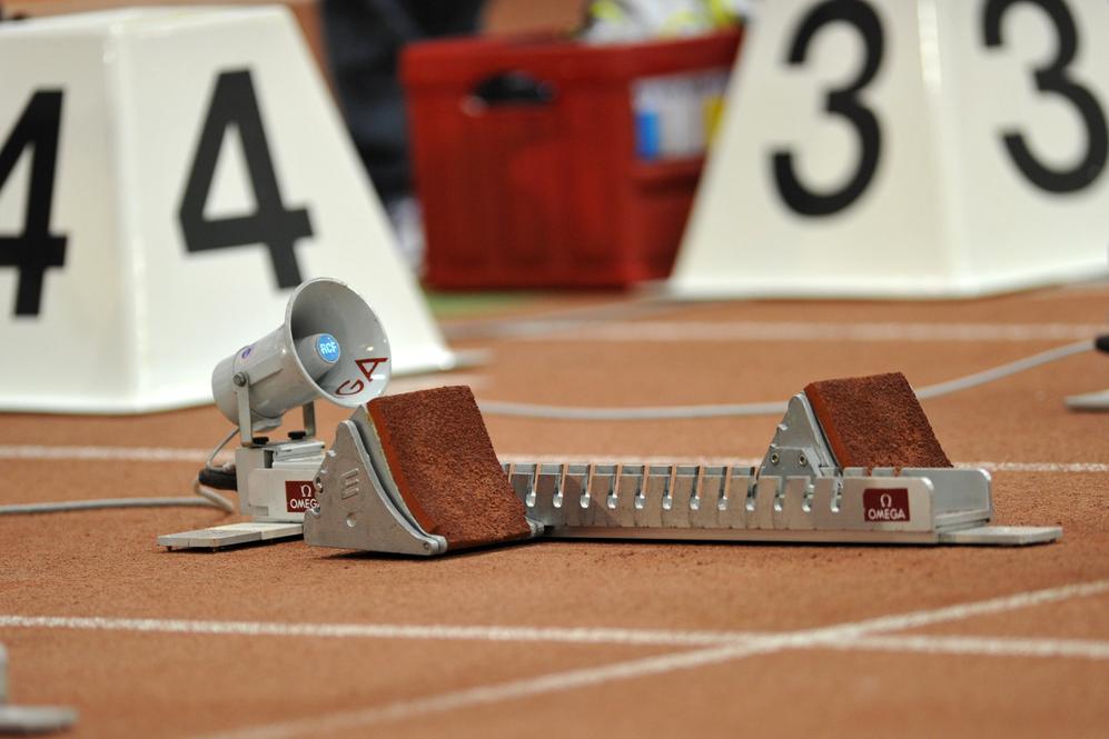 Startblöcke und Zeitmessgeräte auf einer Leichtathletikbahn mit brauner Oberfläche und großen Wettkampfnummern im Hintergrund.