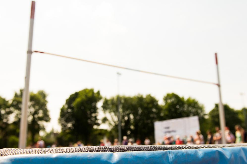 Nahaufnahme einer blauen Sprungmatte mit einer Hochsprunganlage und unscharfem Publikum im Hintergrund.