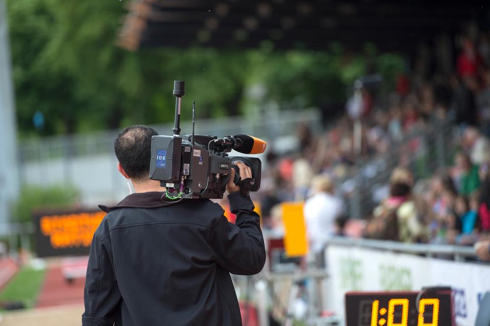 Kameramann filmt bei einer Sportveranstaltung, Hintergrund zeigt eine Zuschauertribüne und eine Zeitmessanzeige.