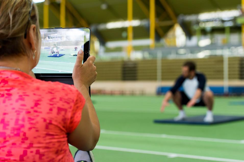 Eine Person filmt einen Mann, der auf einer Matte in der Squat-Position trainiert, in einem Sportzentrum.
