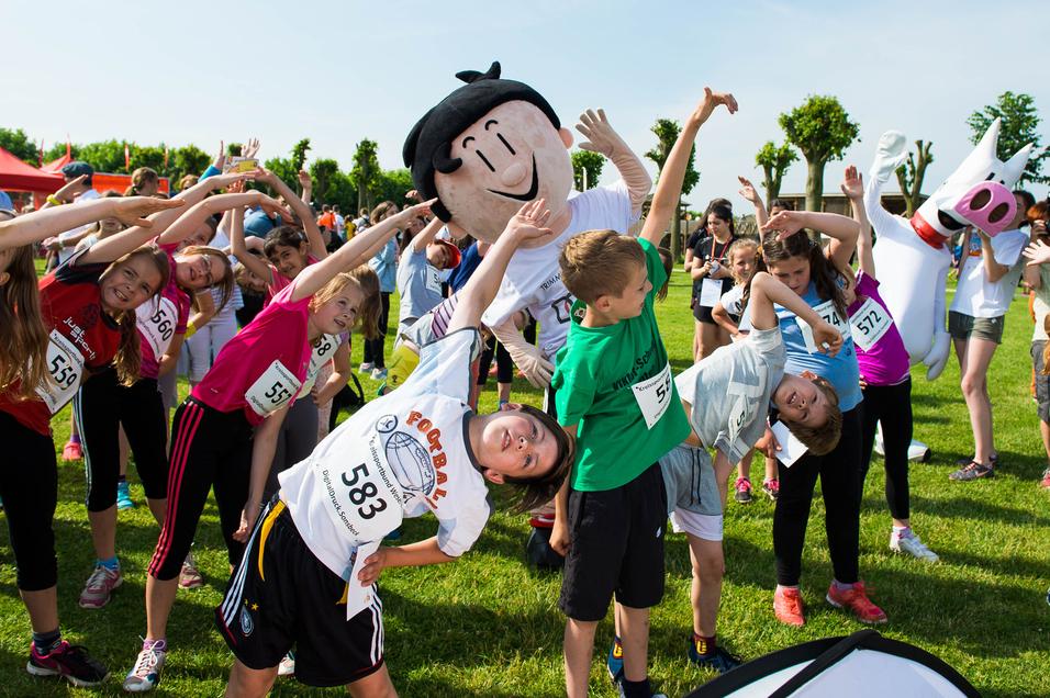 Gruppierung von Kindern in Sportkleidung, die sich gemeinsam dehnen, begleitet von einem großen Maskottchen.