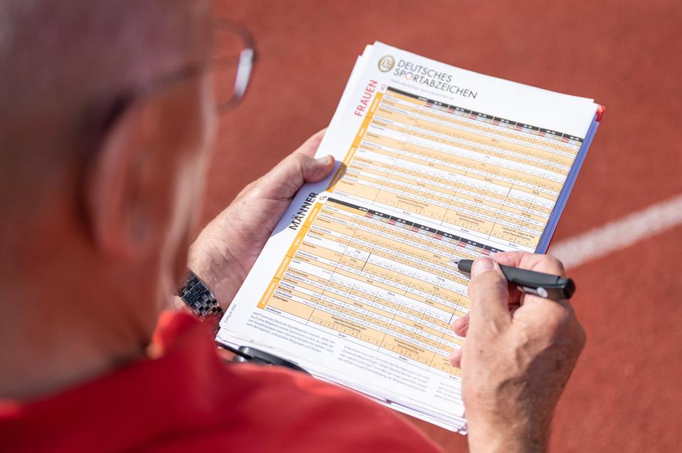 Hand eines Prüfers, der auf einem Blatt mit Sportabzeichen-Anforderungen auf einer Laufbahn notiert.