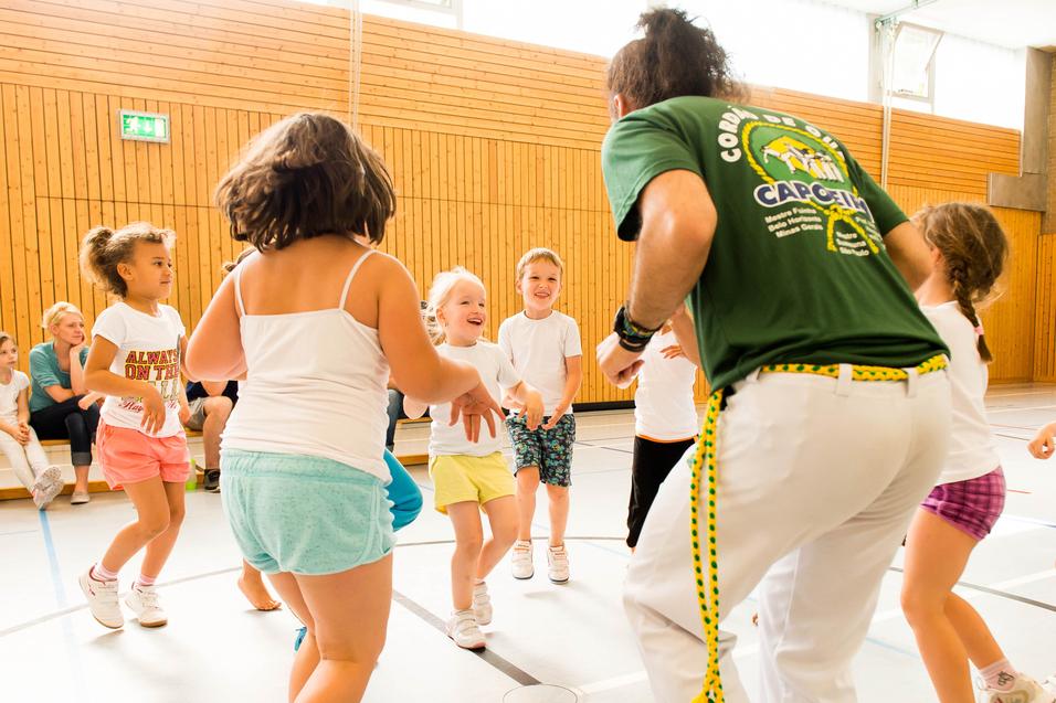 Gruppe von Kindern tanzt fröhlich in einer Turnhalle, geleitet von einem Trainer in grünem T-Shirt.