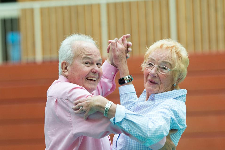 Ein älteres Paar tanzt lächelnd in einer Turnhalle, Kleidung in sanften Farben. Holzpaneele im Hintergrund.