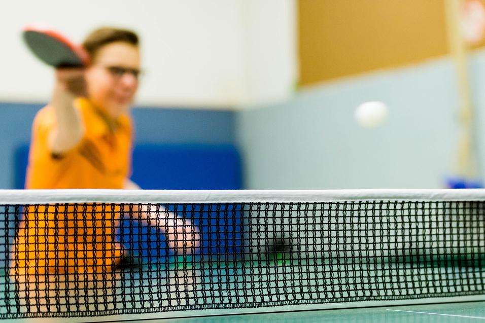 Tischtennisschläger in der Hand eines Spielers, im Hintergrund ein Ball über das Netz fliegend auf einer grünen Tischtennisplatte.