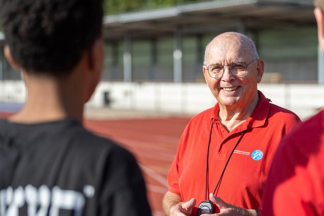 Ein älterer Mann in rotem Shirt mit Stoppuhr spricht an einer Laufbahn mit zwei Jugendlichen.